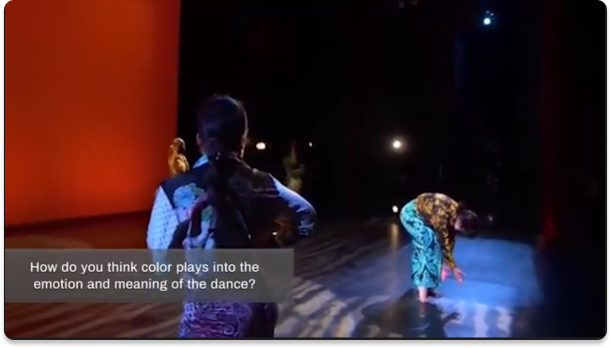 A screenshot of a Kelly Strayhorn Theater dance performance with a overlay at the bottom that says 'How do you think color plays into the emotion and meaning of the dance?'. There are three dancers and one is in the foreground standing tall, one is on the right folded over, and one is in the background out of sight. The background is red and the dancers have colorful blue and yellow clothing.