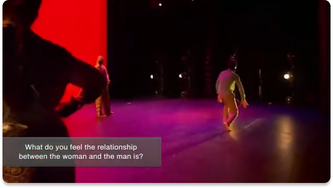 A screenshot of a Kelly Strayhorn Theater dance performance with a overlay at the bottom that says 'What do you think is the relationship between the man and the woman?'. There are three dancers and one is in the foreground standing tall, one is on the right crouching away from the first and having their hands outward close to their body, and one is in the background farther away from the camera also standing tall. The background is red and the dancers have colorful blue and yellow clothing. The lighting is pink.